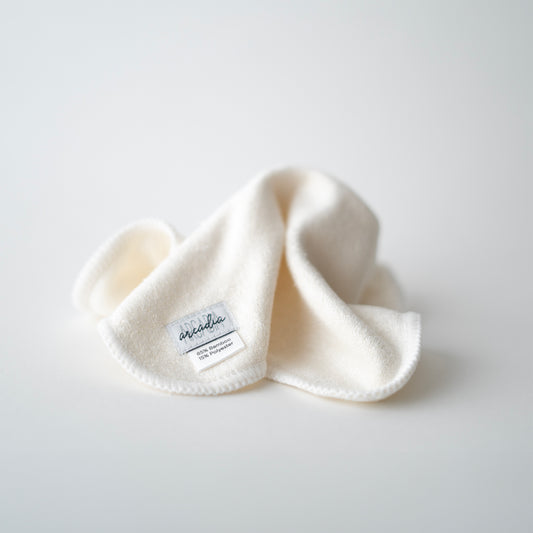 A reusable cloth wipe, which is white with a gentle texture. A small fabric label is stitched to one corner, reading "Arcadia" in a stylish script, along with the text "85% Bamboo, 15% Polyester." The wipe is loosely folded, creating soft shadows that highlight its luxurious and eco friendly appearance.