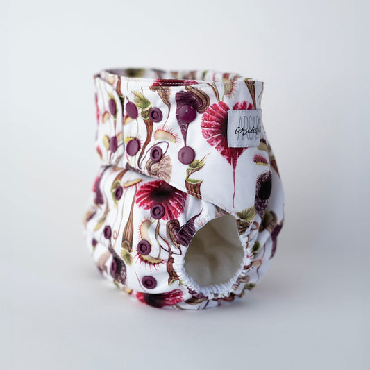 Standing cloth diaper displayed upright to show its structure, with leg openings and adjustable deep plum snaps. The fabric features a bold and bright design of venus flytraps and other carnivorous plants in red, purple, green, and brown hues with a white background. A sewn Arcadia's Nursery logo tag is visible on the front. Front angle view.