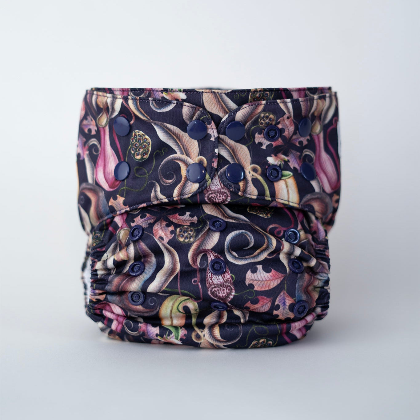 A standing cloth diaper is displayed against a clean white background. The fabric showcases a striking print of pitcher plants, leaves, and seed pods on a purple/blue base. Coordinating deep purple-blue snaps complement the bold, carnivorous plant design. A large amount of snaps on the wing of the diaper and body allow for greater adjustability.