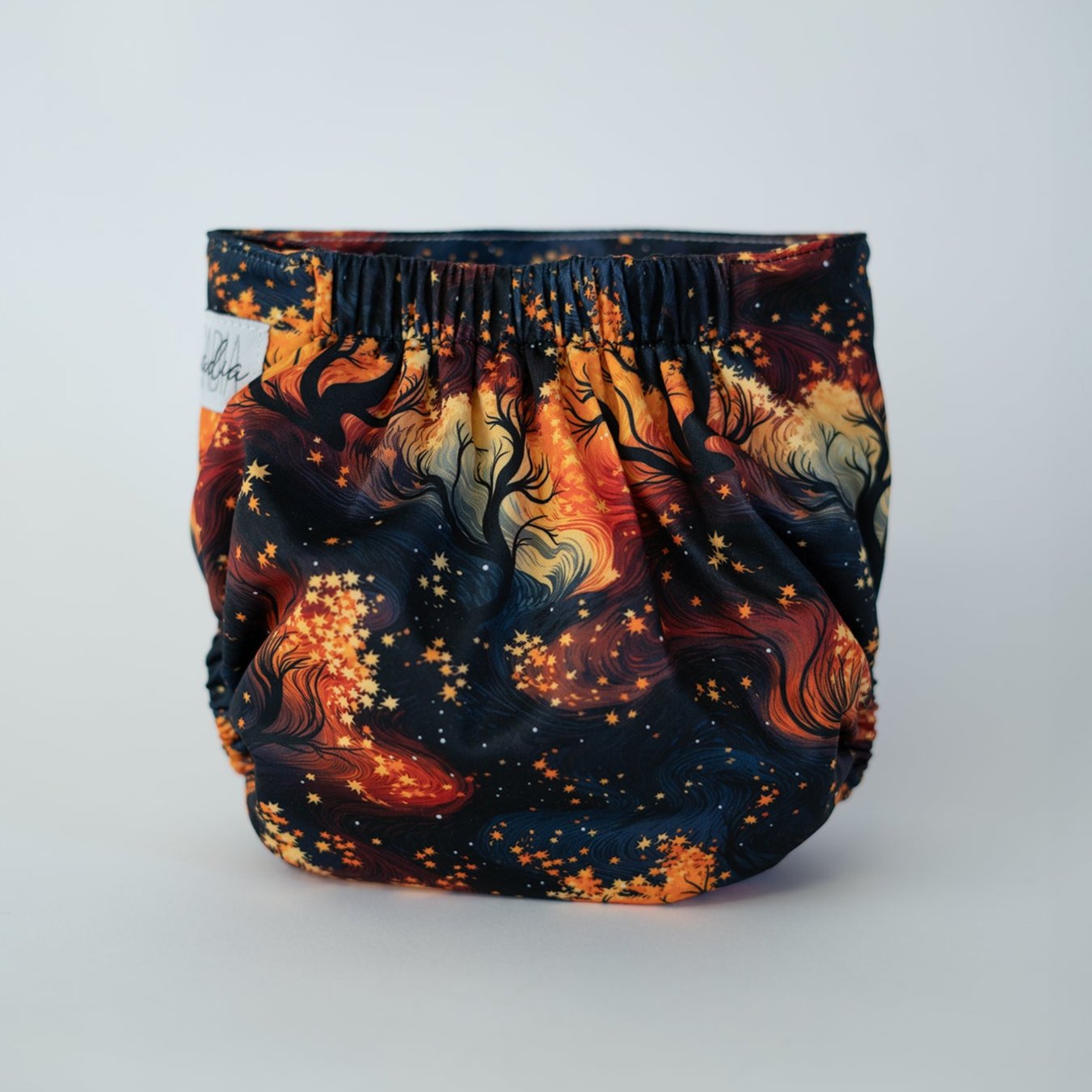 The back view of a pocket style cloth diaper in the Autumn print, photographed against a clean white background. The design is a vibrant fall print, featuring a swirling array of trees, fiery oranges, radiant yellows, depicting leaves falling from the trees. The gathered elastic waistband creates a comfortable, secure fit