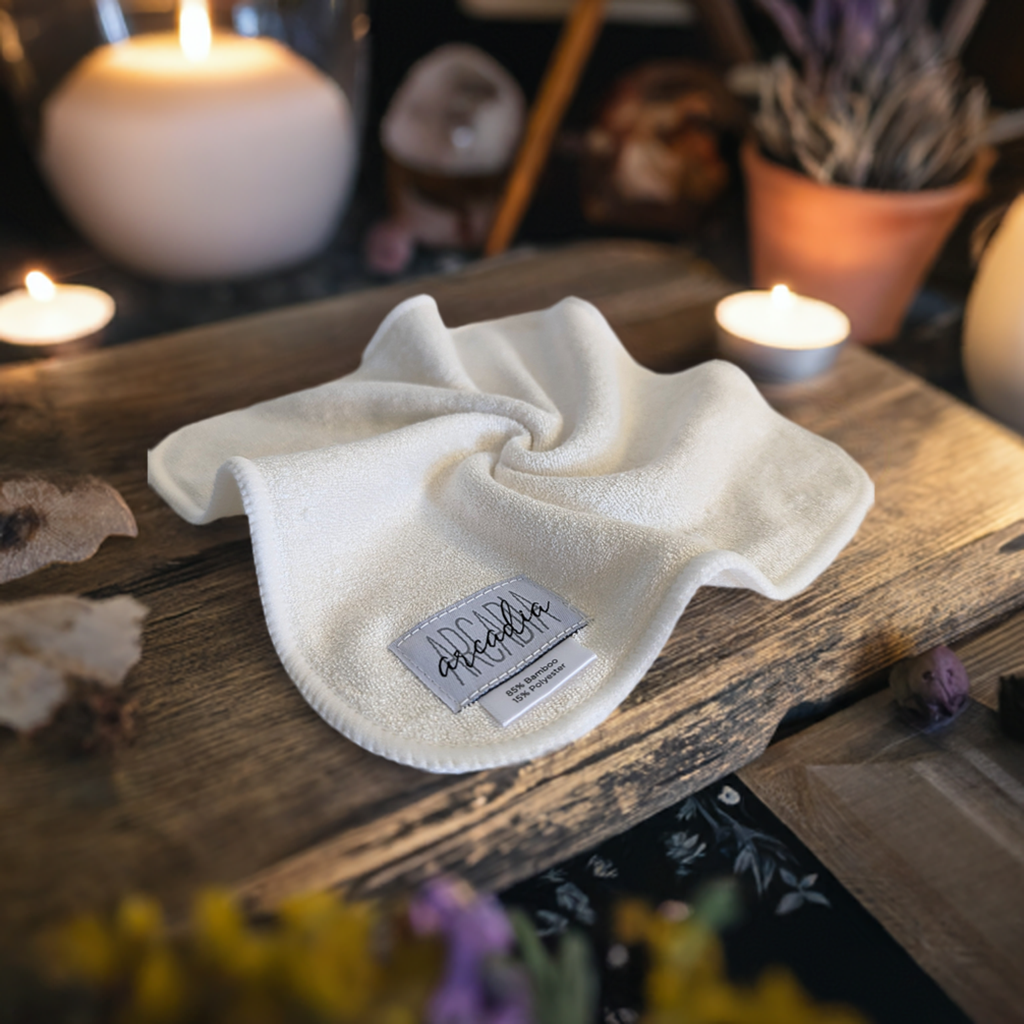 This styled product photo features a reusable cloth wipe laid on a rustic wooden surface. The wipe is white with a soft, plush texture. A fabric label is sewn into one corner, displaying the brand name Arcadia, and noting the material composition: "85% Bamboo, 15% Polyester." The scene is warmly lit, with glowing candles in the background, creating a cozy, inviting atmosphere. Surrounding elements, such as dried flowers and pottery.