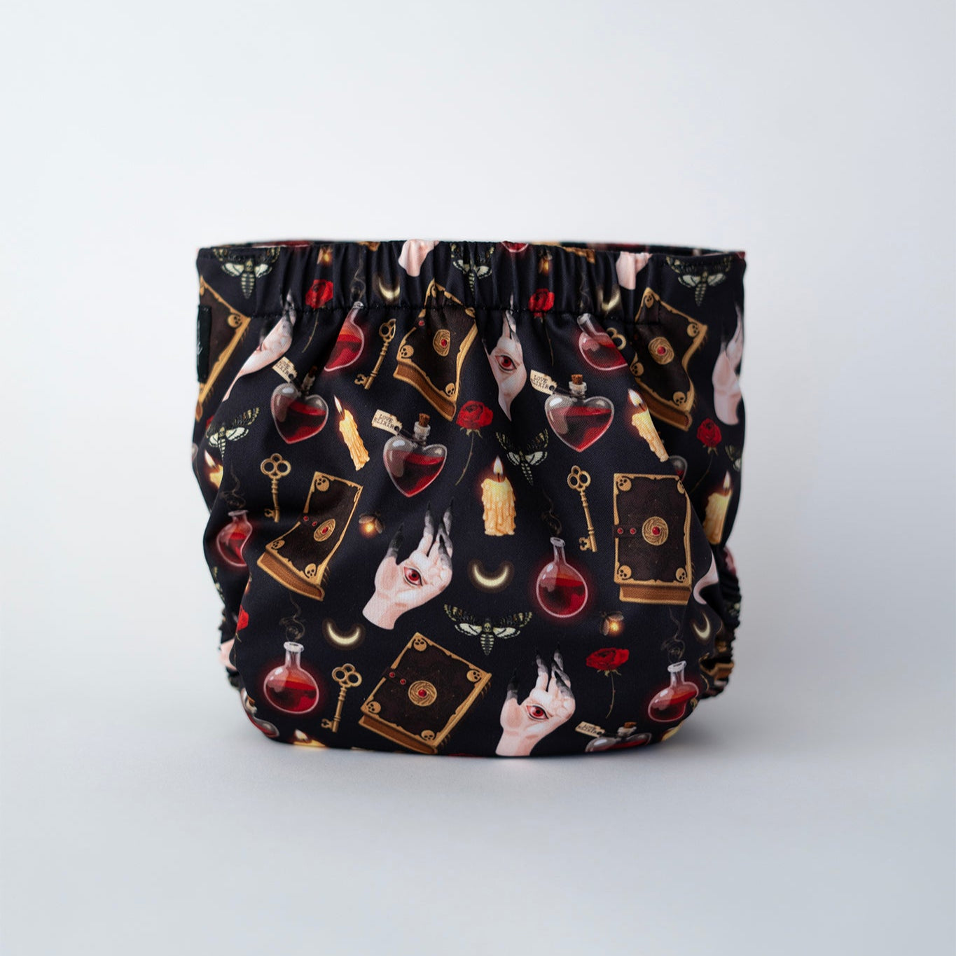 This pocket cloth diaper print features a dark, enchanting design with such as potion bottles, spell books, roses, candles, crescent moons, keys, and mystical symbols against a black background. Back view.