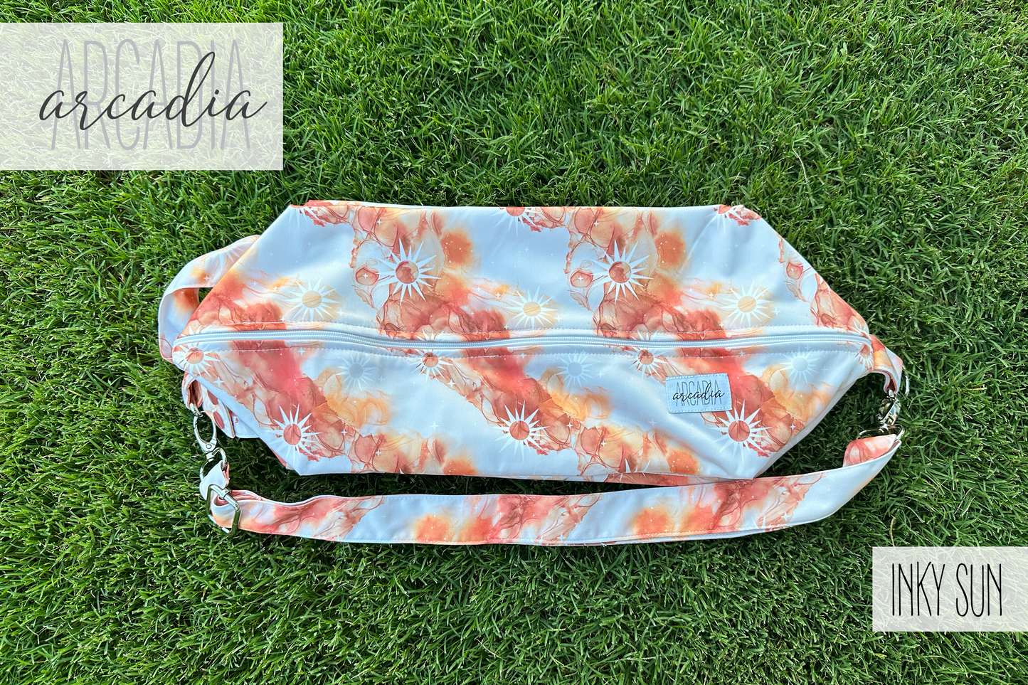 The "Inky Sun" patterned cloth diaper pod, with its orange, rust, and white abstract sun design, is lying flat on vibrant green grass. The adjustable carrying strap is visible, along with silver hardware. The top zipper closure is visible. In the top left corner, the word "arcadia" is written in a script font. In the bottom right corner, the words "INKY SUN" are in a block font. The scene is illuminated by natural daylight.