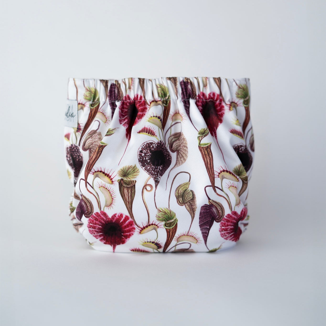 Cloth diaper standing up against a white background, featuring a daring botanical print of carnivorous plants like Venus flytraps and pitcher plants in shades of burgundy, green, and brown on a crisp white fabric. The elastic gathers along the top edge, showcasing the roomy interior and the vivid pattern design. Back view.