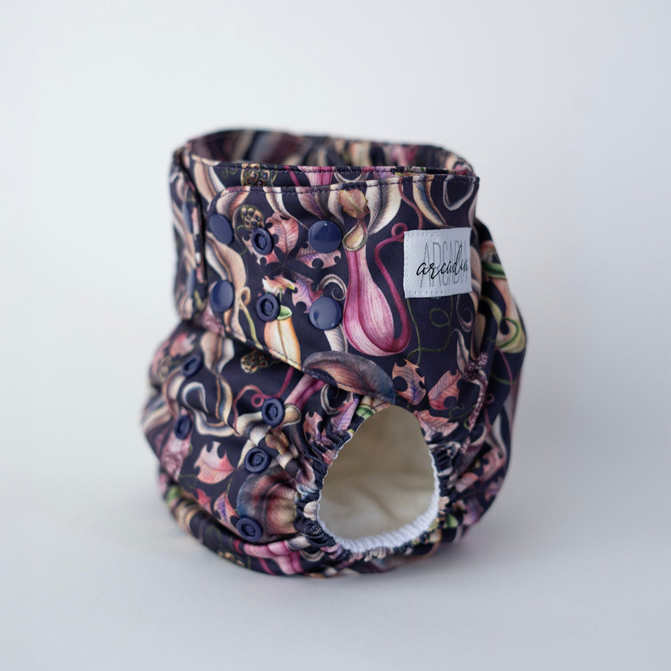 A photo of a cloth diaper standing up against a clean white backdrop. The print features bold pitcher plants, leaves, and seed pods on a deep purple-blue background. Dark purple/blue snaps coordinate with the carnivorous print. You can see the white label sewn to the side of the tab, and the leg boasts comfortable elastics. Front/side angle view.