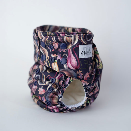 A photo of a cloth diaper standing up against a clean white backdrop. The print features bold pitcher plants, leaves, and seed pods on a deep purple-blue background. Dark purple/blue snaps coordinate with the carnivorous print. You can see the white label sewn to the side of the tab, and the leg boasts comfortable elastics. Front/side angle view.