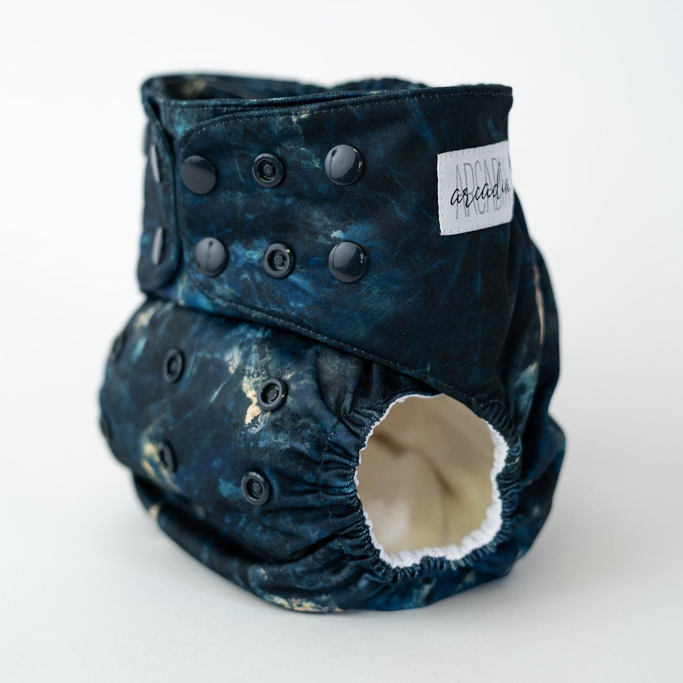 A pocket style cloth diaper from Arcadia’s Nursery shown at a slight front angle in the Apatite print. The fabric displays rich blues, gold veins/splotches, to give the look of a moody crystal. Coordinating deep blue snap closures along the waist and rise provide an adjustable fit, and the curved leg openings highlight the performance focused design.