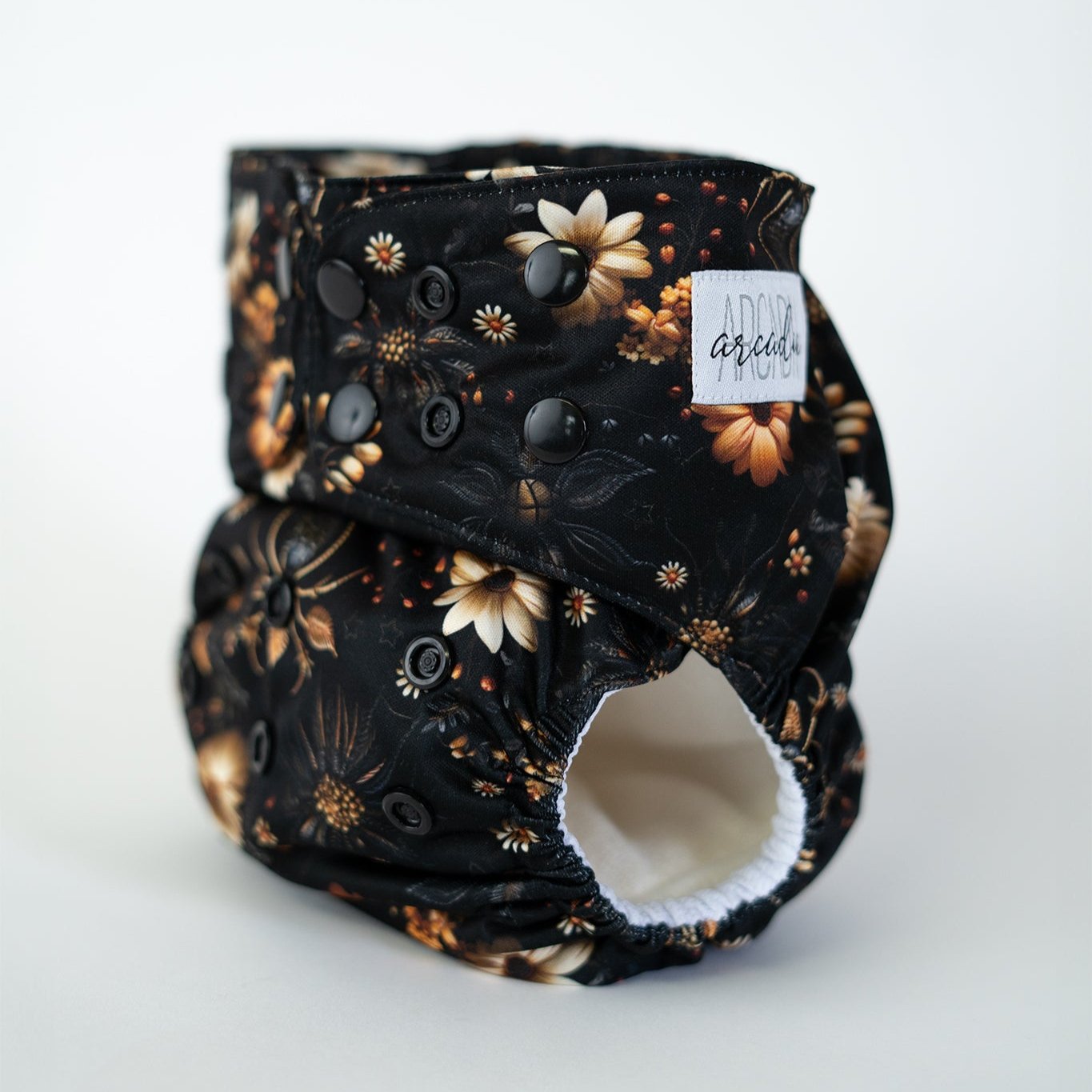 A pocket cloth diaper in the Arachniflora print shown at a front angle, displaying adjustable black snaps along the waist and rise. The print combines ornate florals and carefully illustrated spiders in a dimensional faux stitched style, giving the illusion of embroidered detail. The dark background enhances the golden and ivory blooms, creating a moody, botanical design with a bold, gothic edge. An Arcadia's Nursery logo tag is stitched along the side of the tab on the waist.