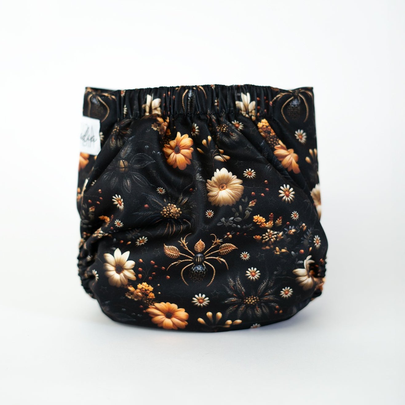 The back view of a pocket style cloth diaper in the Arachniflora print, set against a white background. The print features spiders nestled among warm toned florals in a detailed 3D faux embroidery style. Golden blossoms, delicate leaves, and subtle web motifs stand out against a deep black background, while the gathered elastic waistband is visible at the top of the back.