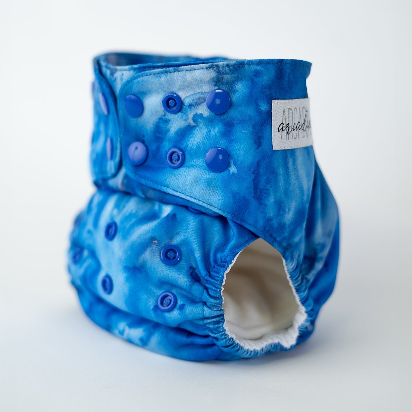 A photo of a vibrant, watercolor blue cloth diaper displayed against a clean, white background. The diaper features an elastic waistband designed to keep your baby comfortable. This modern cloth diaper adds a touch of style with its bold, watercolor-like pattern, making it a perfect choice for parents who value both function and fashion. Front angle view.