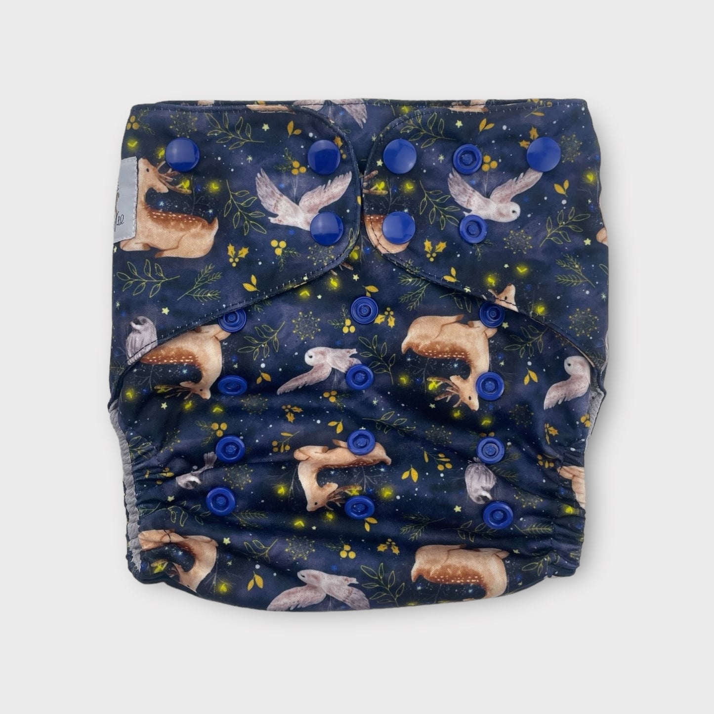 A pocket style cloth diaper with a dark navy background and illustrations of owls, deer, fireflies, and minimalist foliage in yellow. the snaps are dark blue.