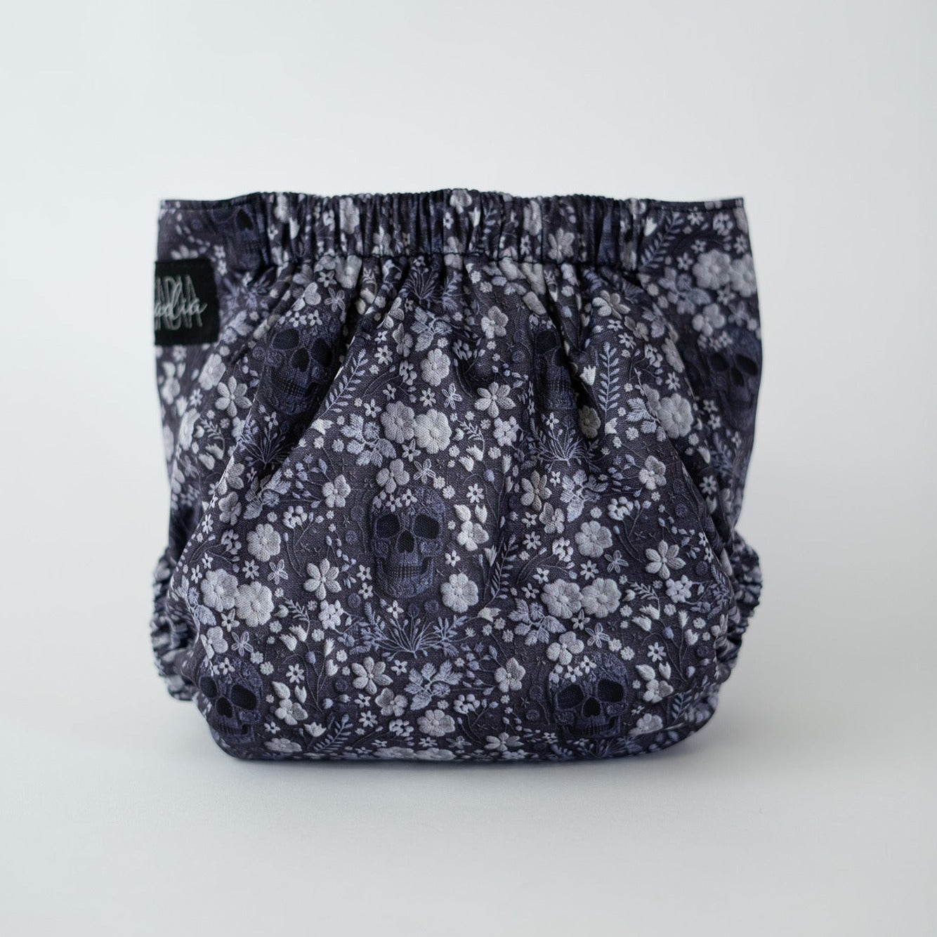 The back view of a pocket style cloth diaper in The Bones & Blooms print from Arcadia’s Nursery. Flowers, skulls, and feathers repeat across the fabric in shades of charcoal, black, and gray in a faux embroiders Soft elastic at the waist and legs creates a contoured shape, while the uninterrupted pattern emphasizes the dark, alternative style.