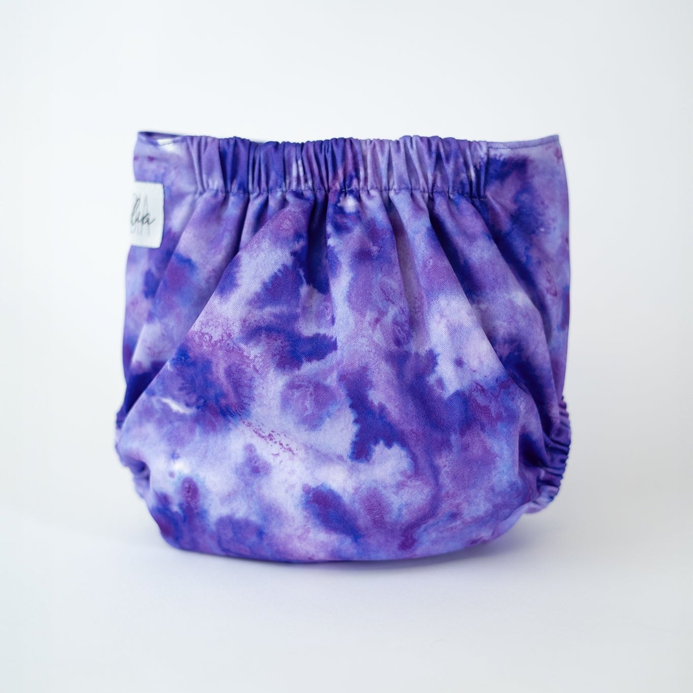 A shot of a vibrant, tie dye printed purple cloth diaper displayed against a clean, white background. The diaper features an elastic waistband for a snug fit designed to keep your baby comfortable. This modern cloth diaper adds a touch of style with its bold, watercolor pattern, making it a perfect choice for parents who value both function and fashion. Rear view.