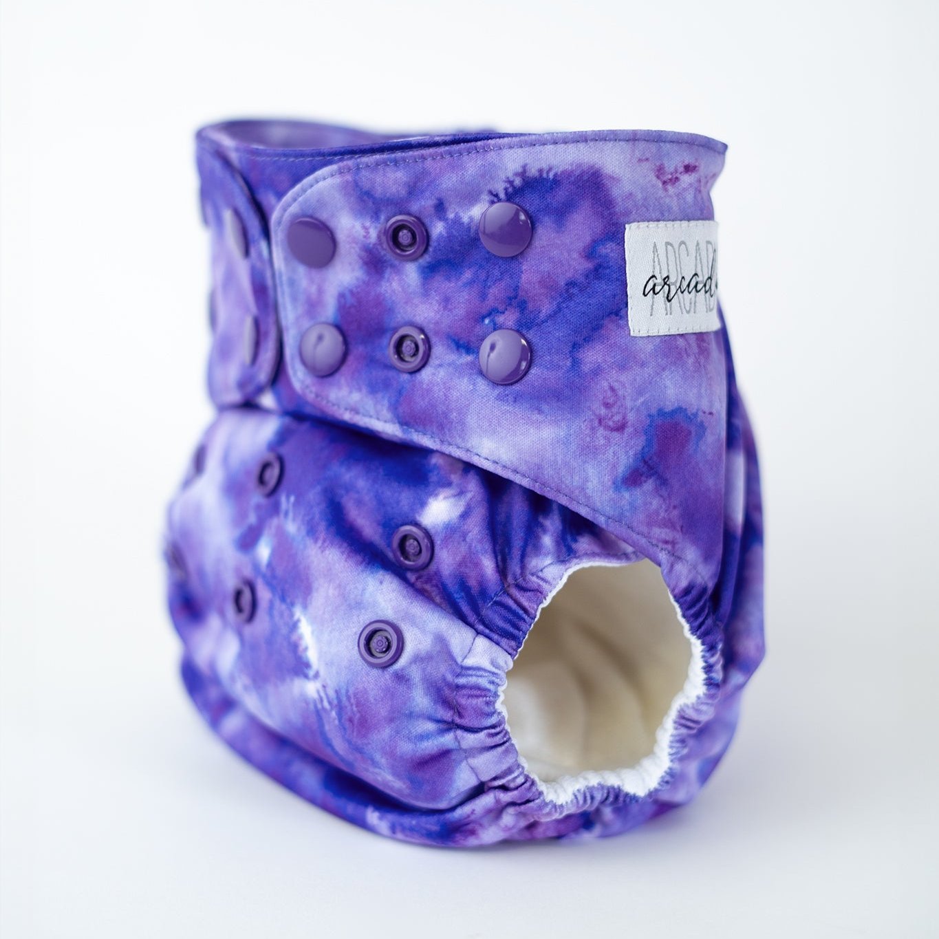 A shot of a vibrant, tie dye printed purple cloth diaper displayed against a clean, white background. The diaper features an elastic waistband for a snug fit designed to keep your baby comfortable. This modern cloth diaper adds a touch of style with its bold, watercolor pattern, making it a perfect choice for parents who value both function and fashion. Front angle view.