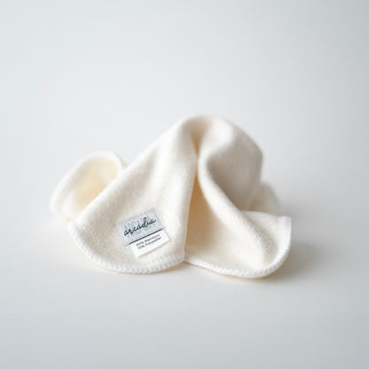 A reusable cloth wipe, which is white with a gentle texture. A small fabric label is stitched to one corner, reading "Arcadia" in a stylish script, along with the text "85% Bamboo, 15% Polyester." The wipe is loosely folded, creating soft shadows that highlight its luxurious and eco friendly appearance.