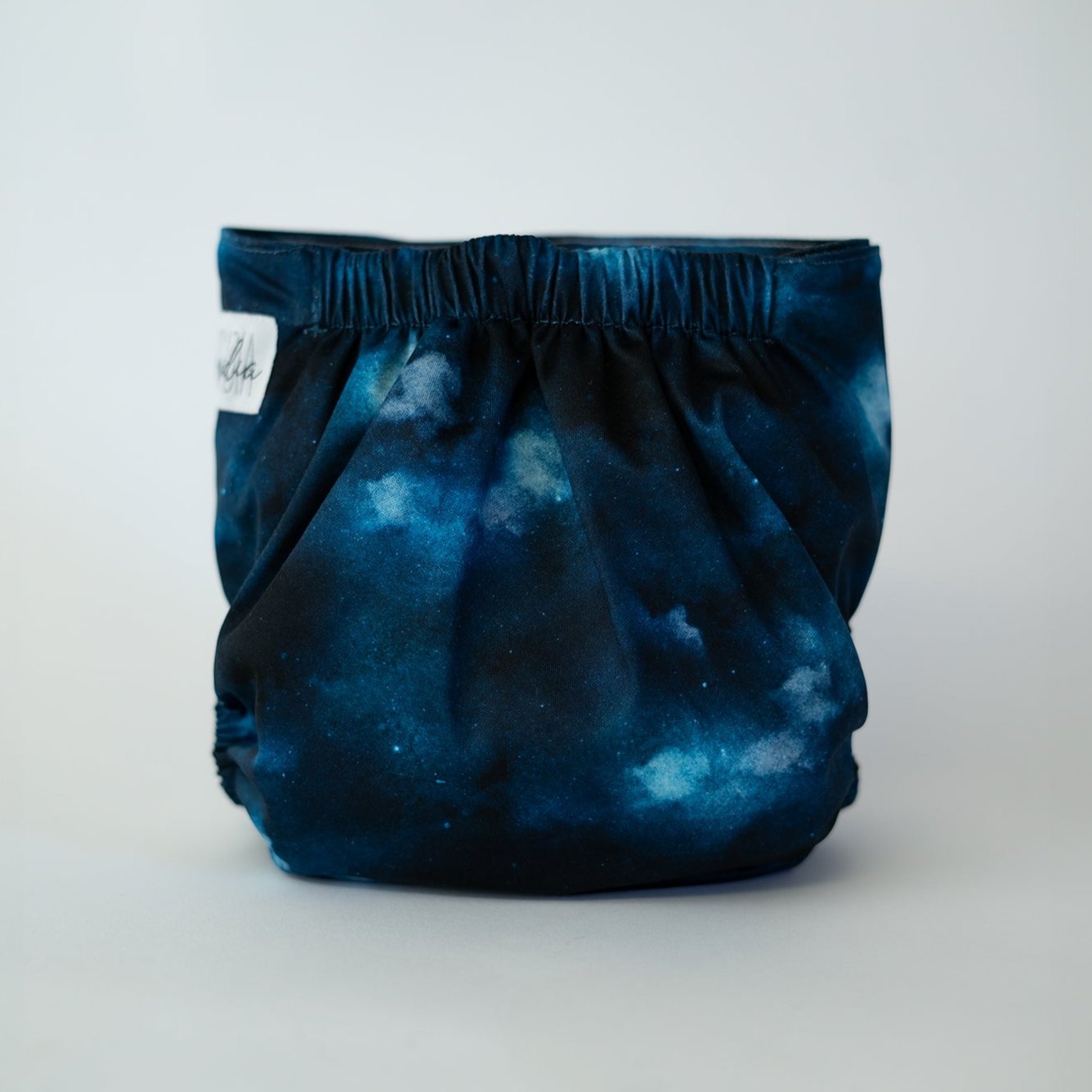 A pocket cloth diaper in the Cosmos print shown from the back. The print shows an ethereal black and blue space scene. The gathered elastic waistband is visible along the top, with the galactic spread across the fabric.