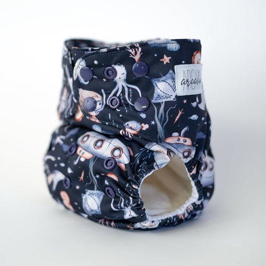 Navy blue pocket cloth diaper featuring an enchanting underwater adventure theme with submarines, narwhals, starfish, and scuba divers in soft pastel colors. There are also octopus, coral, and kelp. This reusable diaper is perfect for parents seeking a fun and imaginative design while ensuring comfort for their little one. Front angle view.