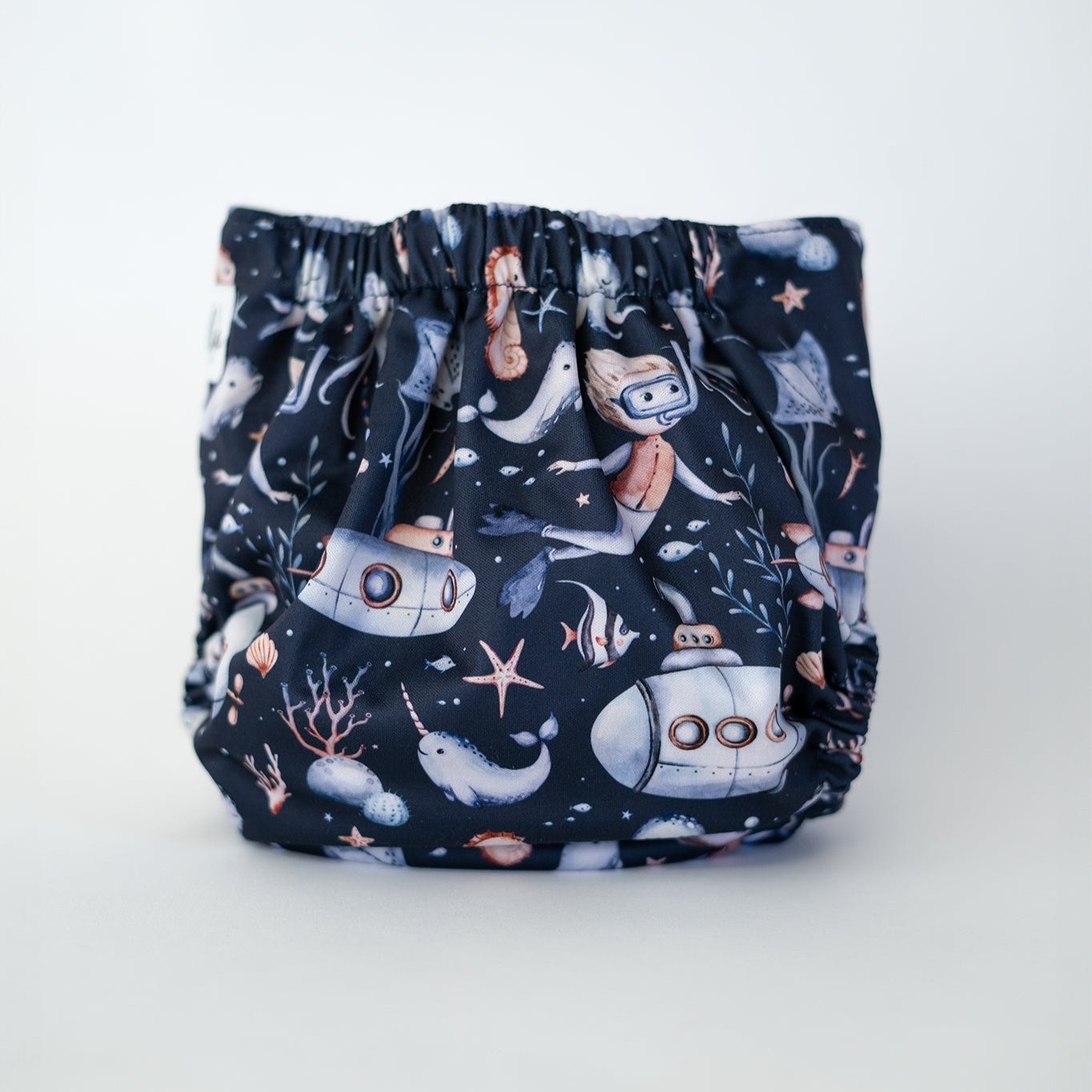 Navy blue pocket cloth diaper featuring an enchanting underwater adventure theme with submarines, narwhals, starfish, and scuba divers in soft pastel colors. There are also octopus, coral, and kelp. This reusable diaper is perfect for parents seeking a fun and imaginative design while ensuring comfort for their little one. Rear view.