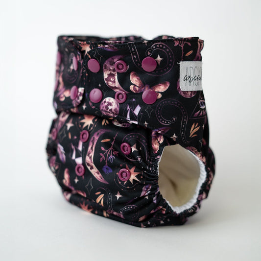 Black pocket cloth diaper featuring a mystical design with moons, stars, crystals, and geometric symbols in shades of purple and pink. This eco-conscious diaper offers a snug fit and unique style, perfect for parents seeking both functionality and fashion in their reusable diapering options.