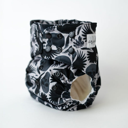 Black and gray pocket cloth diaper with a striking pattern featuring skulls, crows, and feathers against a white background. This eco-friendly diaper is designed for adjustable fit and comfort, combining style with sustainability. Ideal for parents seeking unique, reusable diapering options. Front angle view.