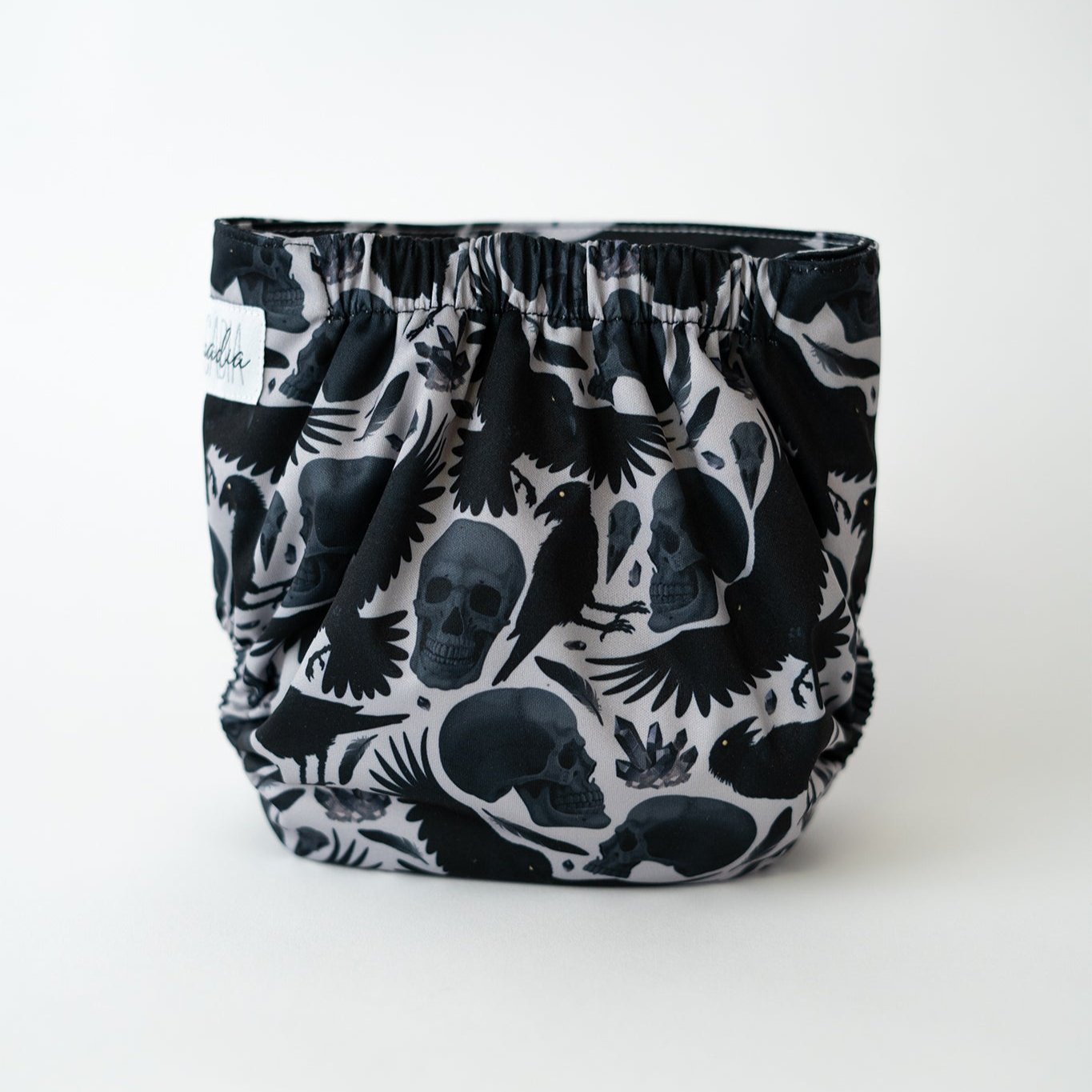 Black and gray pocket cloth diaper with a striking pattern featuring skulls, crows, and feathers against a white background. This eco-friendly diaper is designed for adjustable fit and comfort, combining style with sustainability. Ideal for parents seeking unique, reusable diapering options. Back view.