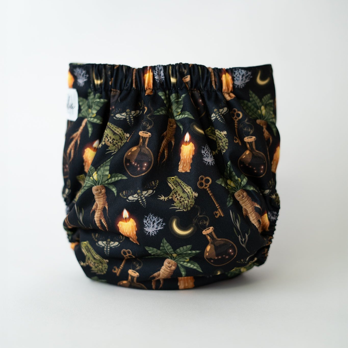 Reusable cloth diaper featuring a witchcraft inspired print. It has spell jars, frogs, moths, mandrakes, keys, and moons. This whimsical, botanical, green witch inspired pattern is perfect for spooky spiritual moms. Rear view.