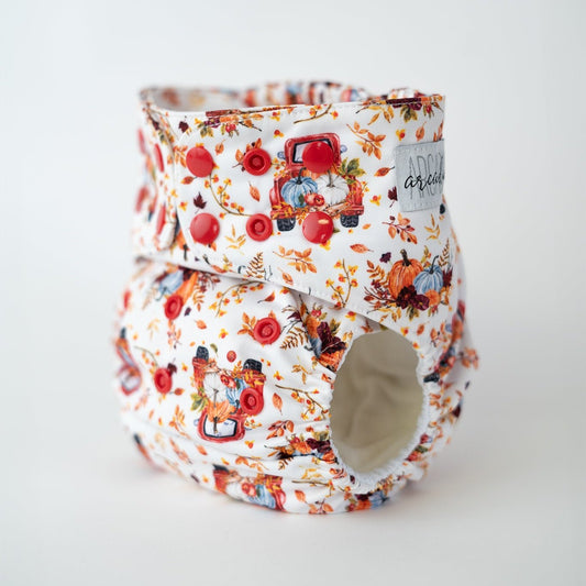 A reusable cloth diaper printed with fall leaves, an orange antique truck, and pumpkins. This fall print features a white background and deep orange snaps.