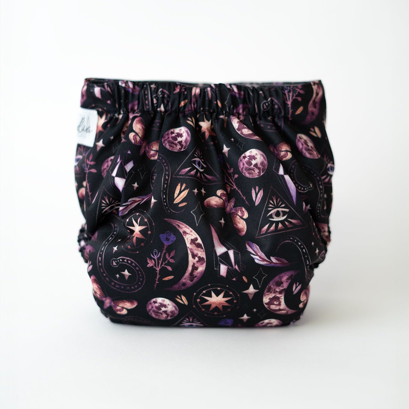 A photo of a pocket cloth diaper with a black background featuring a mystical design with moons, stars, crystals, and geometric symbols in shades of purple and pink. This eco-conscious diaper offers a snug fit and unique style, perfect for parents seeking both functionality and fashion in their reusable diapering options. Rear view.