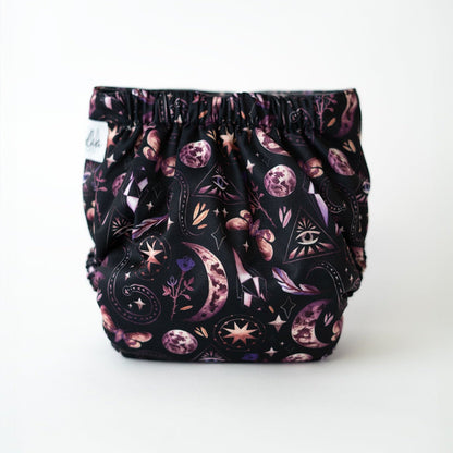 A photo of a pocket cloth diaper with a black background featuring a mystical design with moons, stars, crystals, and geometric symbols in shades of purple and pink. This eco-conscious diaper offers a snug fit and unique style, perfect for parents seeking both functionality and fashion in their reusable diapering options. Rear view.