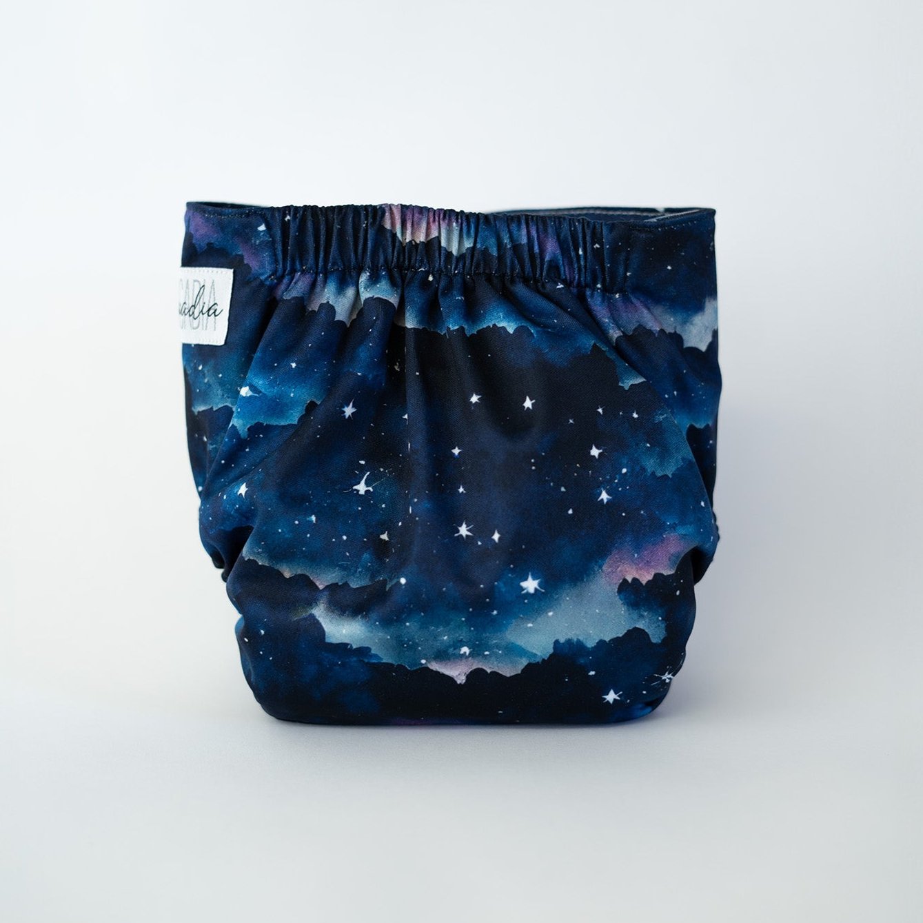 The back view of a pocket cloth diaper in the Dusk print against a white background. The fabric displays a star filled night sky in shades of blue and purple with subtle sunset tones near the horizon and dark silhouetted landscape shapes. The gathered elastic waistband is visible along the top edge.