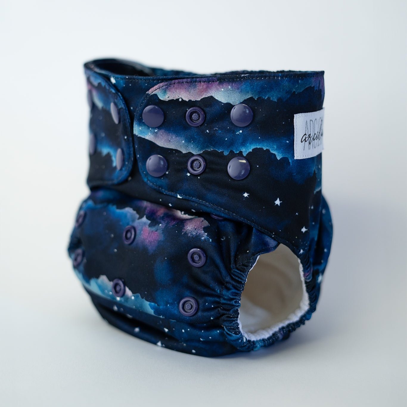 A pocket cloth diaper in the Dusk print shown at a front angle. The fabric features a layered night sky design with deep navy, indigo, and soft purple tones blending like a fading sunset. Small white stars are scattered across the sky above dark landscape silhouettes. Adjustable navy snaps line the waist and rise. The inner lining is white AWJ and visible at the leg opening.