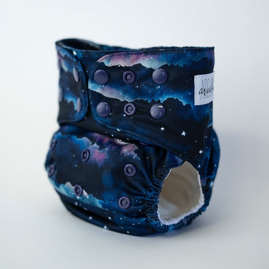 A pocket cloth diaper in the Dusk print shown at a front angle. The fabric features a layered night sky design with deep navy, indigo, and soft purple tones blending like a fading sunset. Small white stars are scattered across the sky above dark landscape silhouettes. Adjustable navy snaps line the waist and rise. The inner lining is white AWJ and visible at the leg opening.