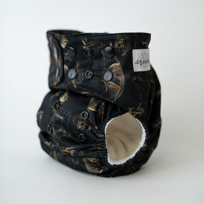 A pocket cloth diaper in the Fig 10 print shown at a front angle. The black fabric features sepia anatomical illustrations including a detailed eye, a jaw in profile, a rib cage, and a heart. Black adjustable snaps are visible along the waist and rise. The leg opening reveals a white AWJ inner lining. An arcadia's nursery logo is sewn into the fabric on the side of the waist.