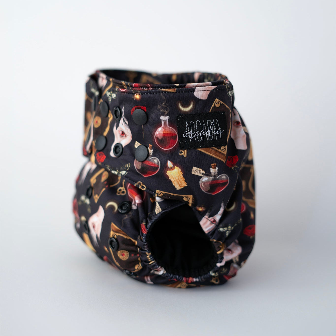 This pocket cloth diaper features a dark, enchanting design with illustrated elements such as potion bottles, candles, crescent moons, keys, spell books, and moths against a black background. Complete with secure black snaps and Arcadia's Nursery branding, this diaper offers a snug and adjustable fit.