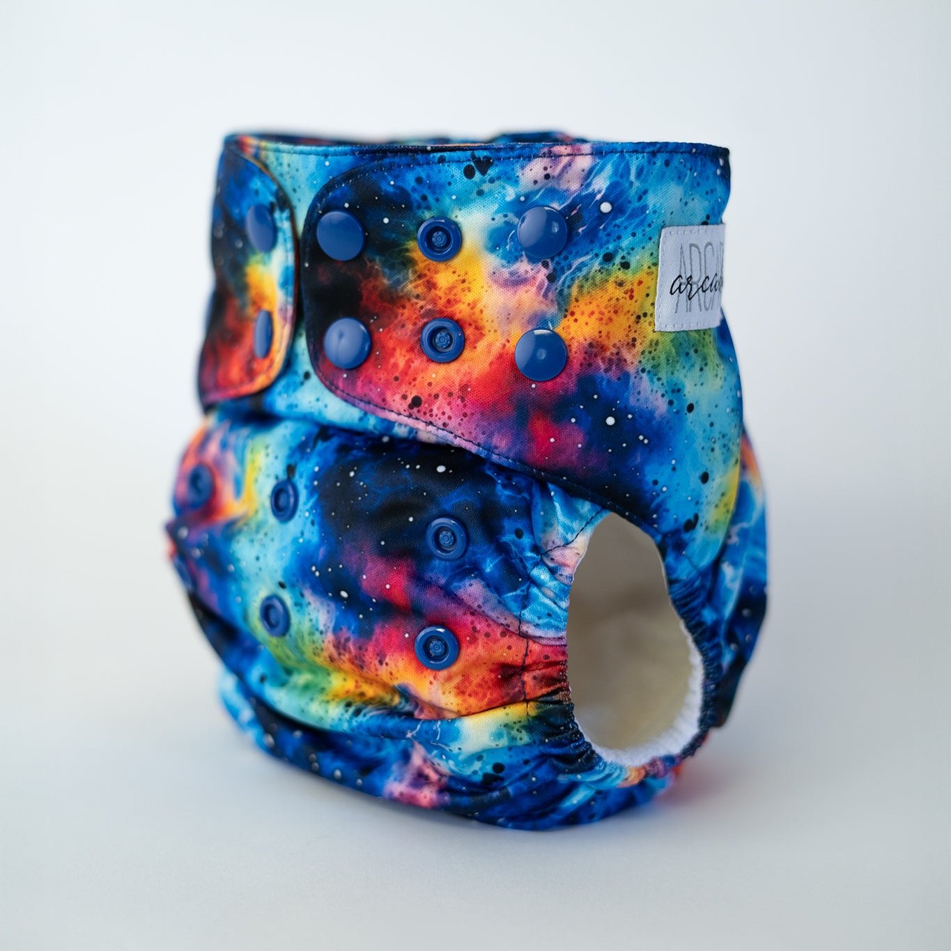A pocket cloth diaper in the Galaxy tie dye print shown at a front angle, revealing rows of coordinating blue adjustable snap closures along the waist and rise. The print showcases a dynamic, fluid alcohol ink pattern of interstellar clouds in electric blues, vivid purples, and bright golds that wrap around the diaper. Contoured leg elastics and a soft white inner lining highlight the functional design, with a small logo tag visible on the wing.