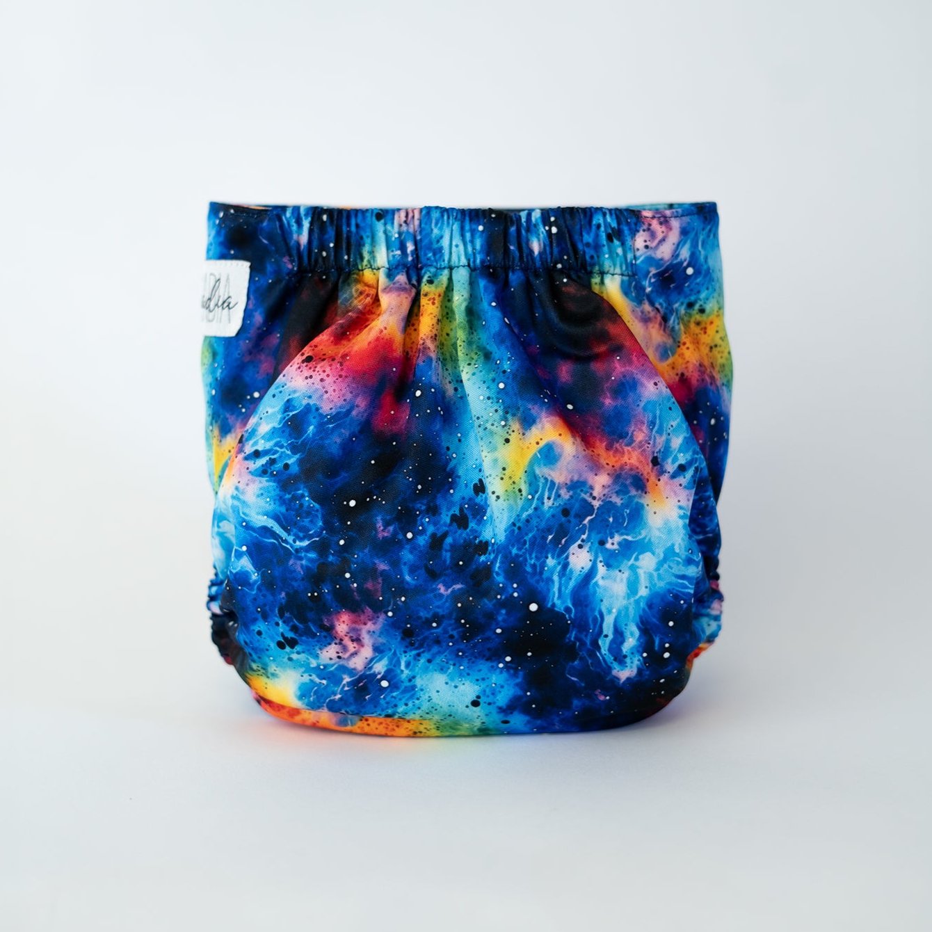 The back view of a pocket style cloth diaper in the Galaxy tie dye print, photographed against a clean white background. The design is a vibrant cosmic watercolor and alcohol ink blend, featuring a swirling galaxy of deep sapphire blues, fiery oranges, radiant yellows, and cosmic magentas that bleed into one another organically. The fabric is dusted with fine white starlight speckles, and the gathered elastic waistband creates a comfortable, secure fit.