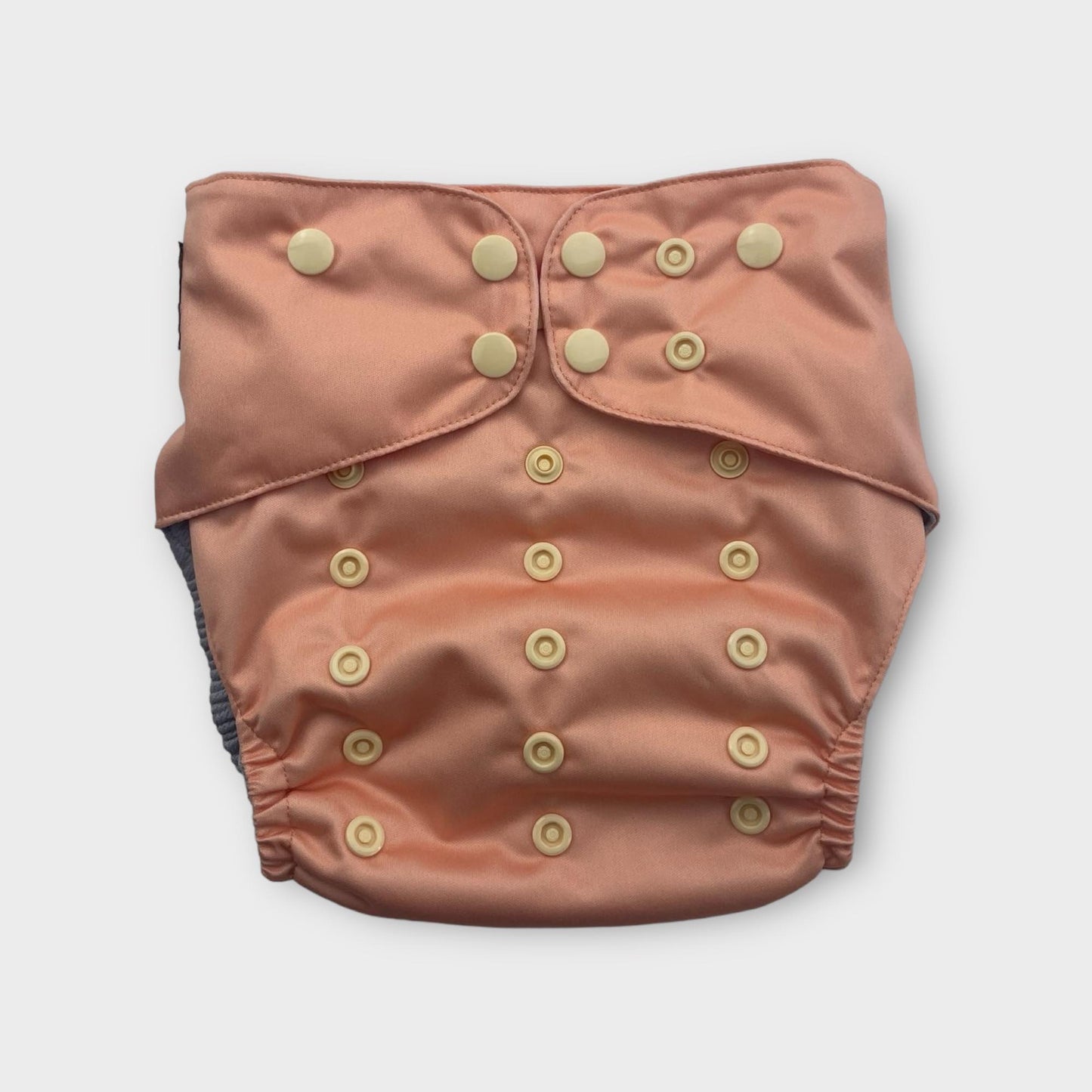 This image shows a front view of a single, reusable cloth diaper in a peach color. It features multiple rows of light-colored snaps on the front, which are used for adjusting the rise and waist size. The diaper has elasticized leg openings, and a dark gray inner lining is visible at the edges. The fabric appears to be soft and slightly lustrous. The diaper is centered on a plain white background under even, bright studio lighting.