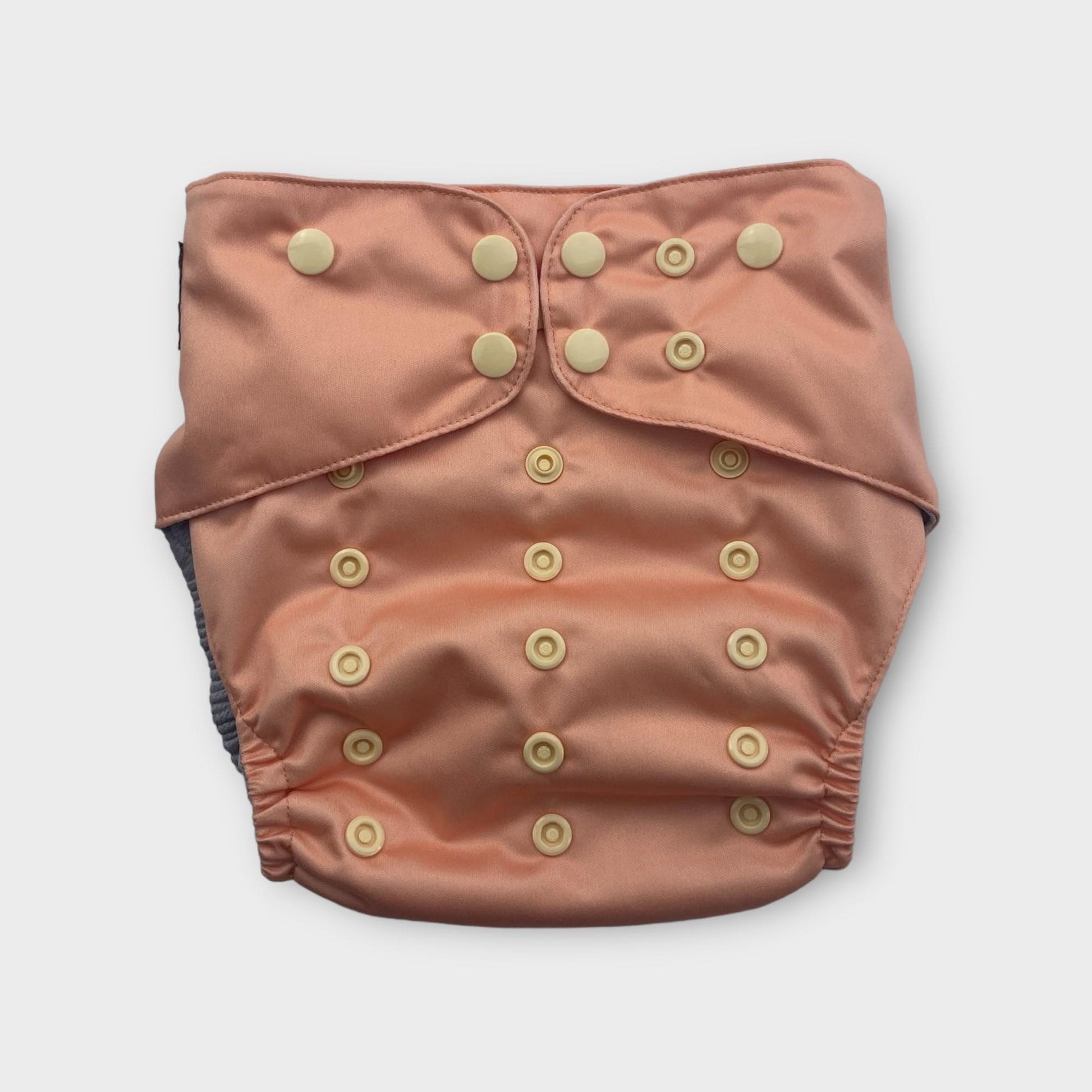 This image shows a front view of a single, reusable cloth diaper in a peach color. It features multiple rows of light-colored snaps on the front, which are used for adjusting the rise and waist size. The diaper has elasticized leg openings, and a dark gray inner lining is visible at the edges. The fabric appears to be soft and slightly lustrous. The diaper is centered on a plain white background under even, bright studio lighting.