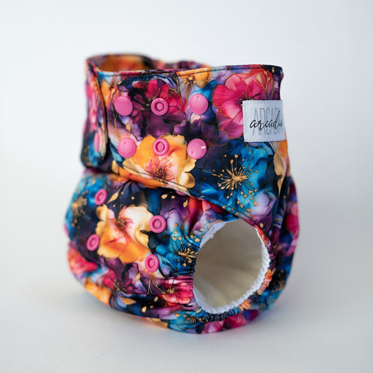 A pocket style cloth diaper from Arcadia’s Nursery shown at a slight front angle in the Gold Vein print. The design features ornate flowers, gold details, in vibrant jewel tones. Adjustable pink snap closures and gathered leg elastics are visible, highlighting the sculpted fit and moody, gothic aesthetic along with white AWJ inside.