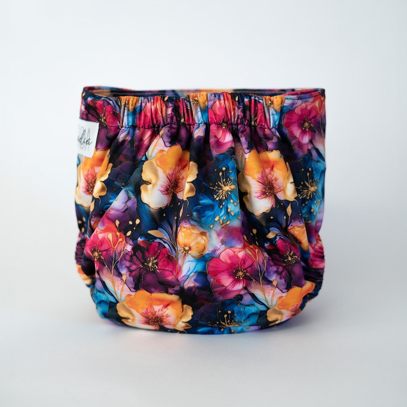 The back view of a pocket style cloth diaper in the Gold Vein print from Arcadia’s Nursery. Stained glass-esque flowers repeat across the fabric in shades of pink, orange, and deep blue with gold details. Soft elastic at the waist and legs creates a contoured shape, while the uninterrupted pattern emphasizes the eccentric and bold.