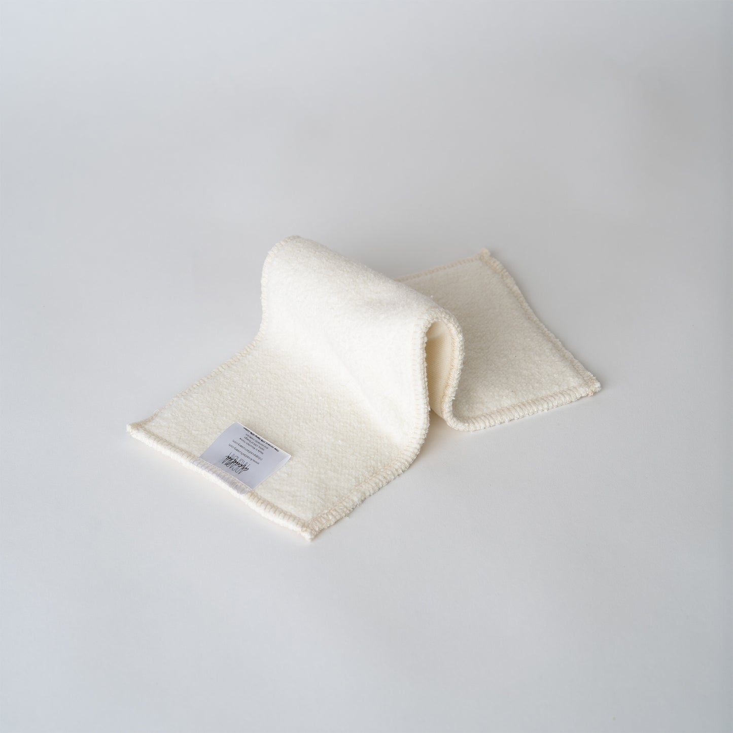 A cream colored cloth diaper insert on a pain white backdrop. the insert is folded in an "inchworm" formation. The insert has squared edges and you can see an information and care tag with the arcadia's nursery logo sewn onto the end.