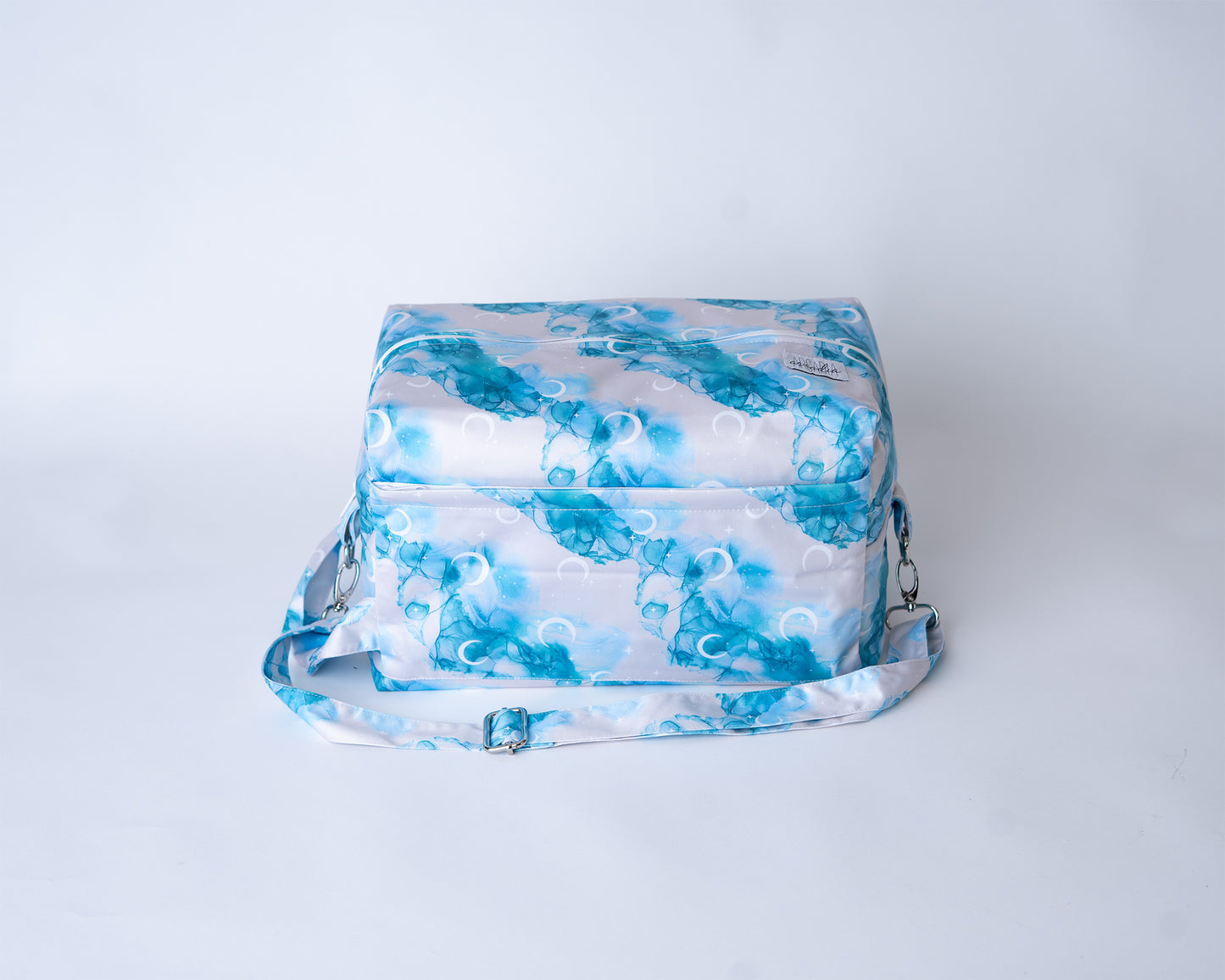 A rectangular cloth diaper pod bag is shown against a plain white studio background. The pod is made from a fabric with the "Inky Moon" print, featuring abstract watercolor designs of moons and alcohol ink swirls in shades of blues, and white on a white background. A matching adjustable shoulder strap with silver metal hardware and clasps is attached to the sides. An Arcadia's logo label is sewn onto the front right corner.