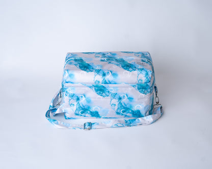 A rectangular cloth diaper pod bag is shown against a plain white studio background. The pod is made from a fabric with the "Inky Moon" print, featuring abstract watercolor designs of moons and alcohol ink swirls in shades of blues, and white on a white background. A matching adjustable shoulder strap with silver metal hardware and clasps is attached to the sides. An Arcadia's logo label is sewn onto the front right corner.