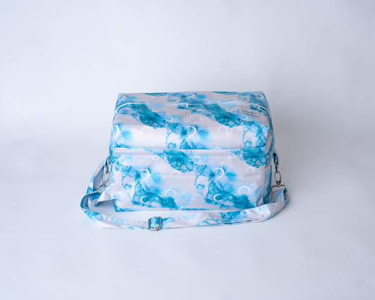 A rectangular cloth diaper pod bag is shown against a plain white studio background. The pod is made from a fabric with the "Inky Moon" print, featuring abstract watercolor designs of moons and alcohol ink swirls in shades of blues, and white on a white background. A matching adjustable shoulder strap with silver metal hardware and clasps is attached to the sides. An Arcadia's logo label is sewn onto the front right corner.