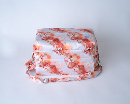 A rectangular cloth diaper pod bag is shown against a plain white studio background. The pod features abstract watercolor designs of suns and swirls in shades of rust, orange, beige, and white on a white/cream background. A matching adjustable shoulder strap with silver metal hardware and clasps is attached to the sides. You can see a front zipper pocket in the photo. An Arcadia's Nursery label is sewn onto the front right corner.