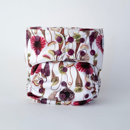Cloth diaper standing up against a white background, featuring a daring botanical print of carnivorous plants like Venus flytraps and pitcher plants in shades of burgundy, green, and brown on a crisp white fabric. The diaper has burgundy snaps. Front view.