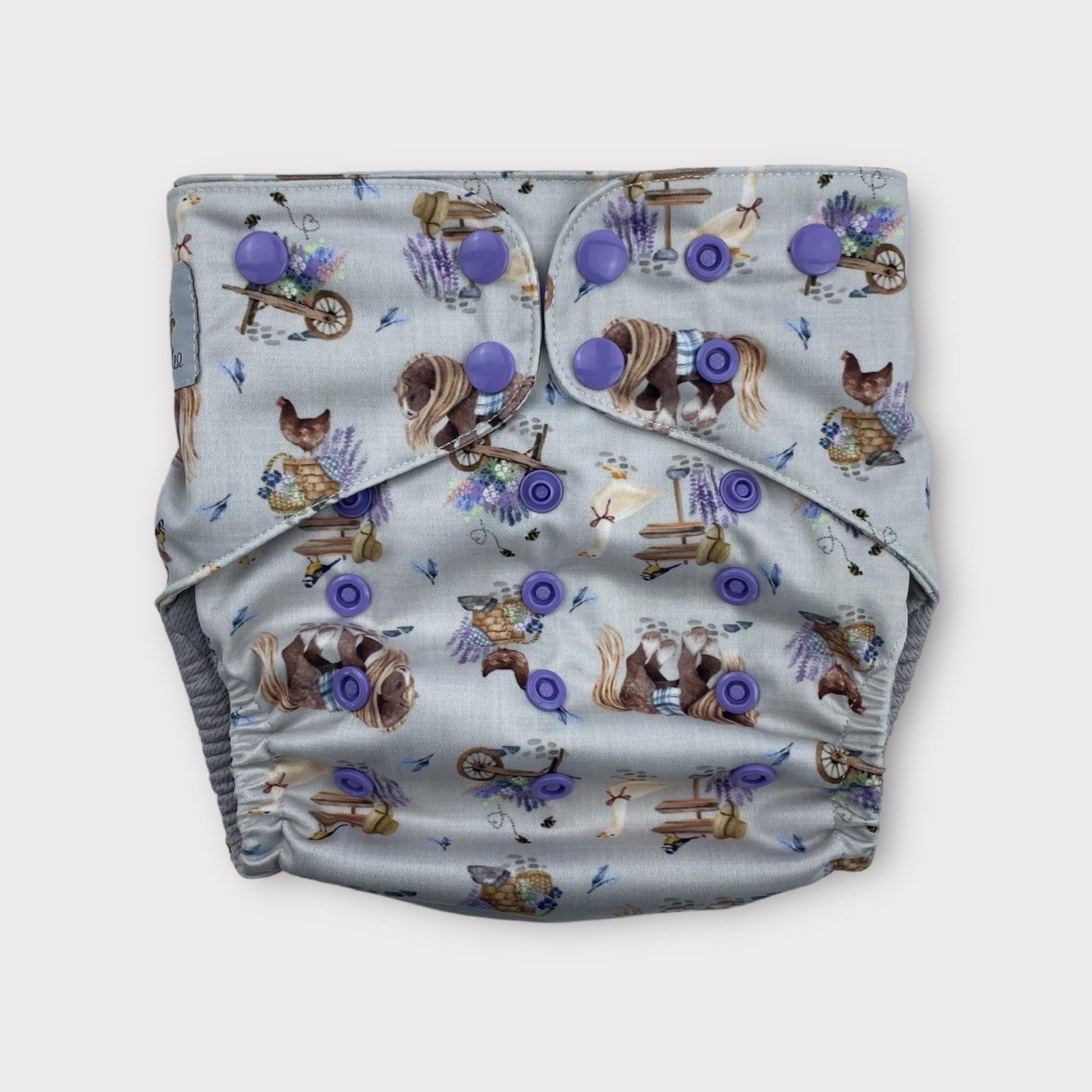 A country cottagecore print wickham wild pocket style reusable cloth diaper with a light grey/blue background and illustrations of chickens, geese, horses, lavender and other floral bouquets, along with signs, baskets and a hat.