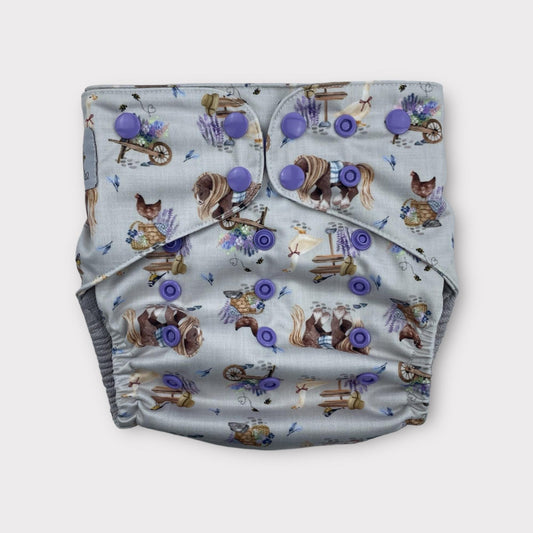 A country cottagecore print wickham wild pocket style reusable cloth diaper with a light grey/blue background and illustrations of chickens, geese, horses, lavender and other floral bouquets, along with signs, baskets and a hat.