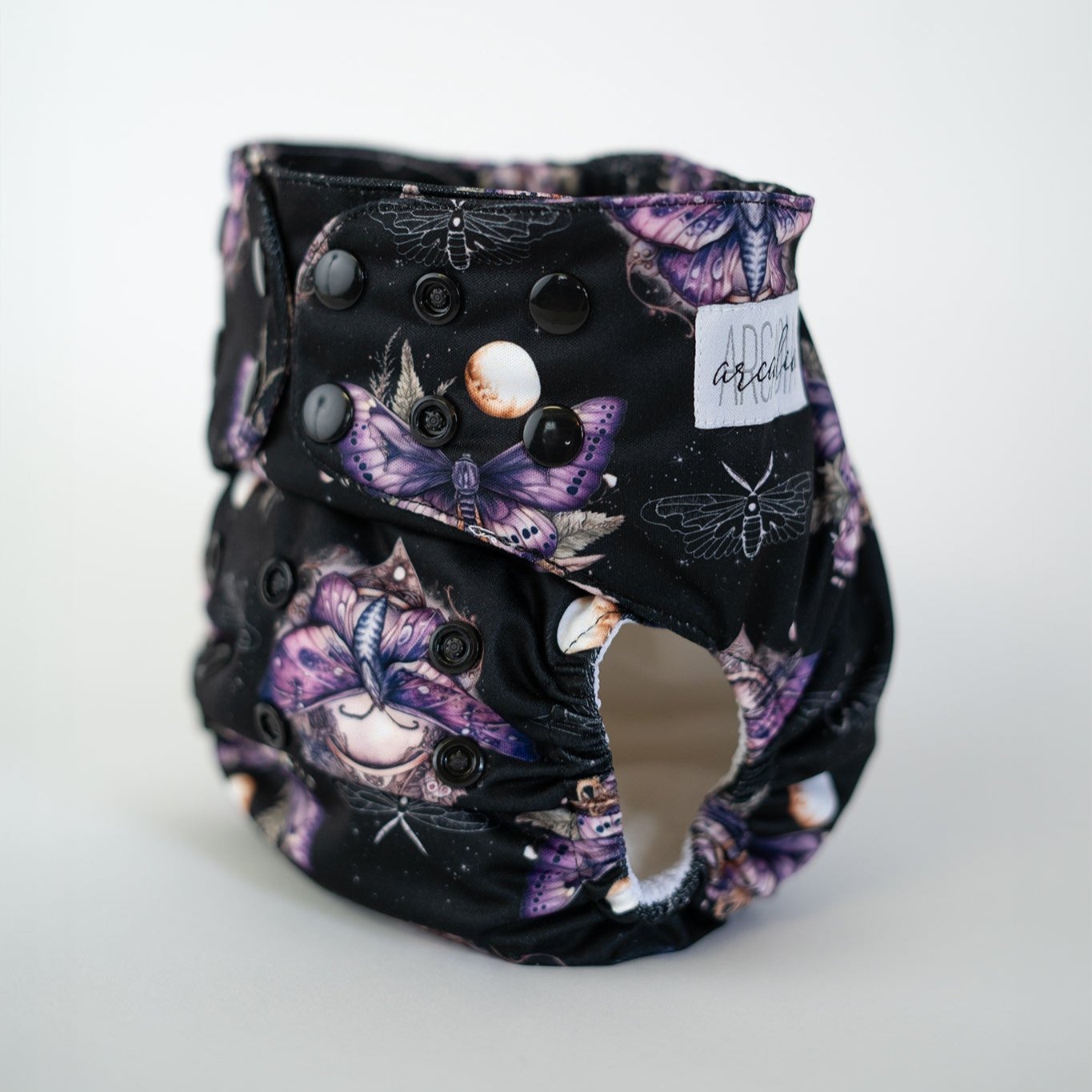 A pocket cloth diaper in the Lepidoptera print shown at a front angle, revealing black adjustable snap closures along the waist & rise. The print showcases an ethereal arrangement of mystical moths & glowing moon icons set against a black background. Contoured leg elastics & a white awj lining highlight the functional design of this otherworldly aesthetic.