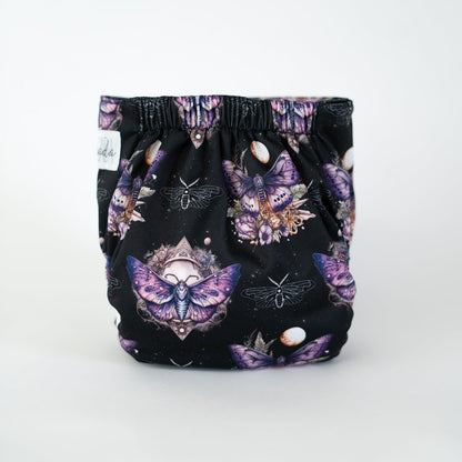 The back view of a cloth diaper in the Lepidoptera print, photographed against a clean white background. The design features a dark, star-dusted backdrop adorned with intricate purple moths, moons, & celestial accents. The gathered elastic waistband provides a snug fit while showcasing the symmetrical, cosmic-inspired artwork that centers on a large, vibrant moth with outstretched wings.